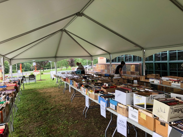 Books for sale on a long table under a tent outside with people browsing